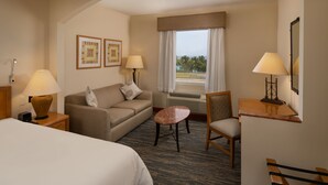 A hotel room with a bed, a sofa, a small table, a chair, and a desk with a lamp. There are framed pictures on the wall and a window with a view. - Four Points by Sheraton Santo Domingo (Boca Chica)