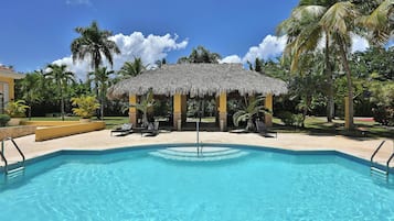 A swimming pool with a thatched gazebo, palm trees, and a clear blue sky.
