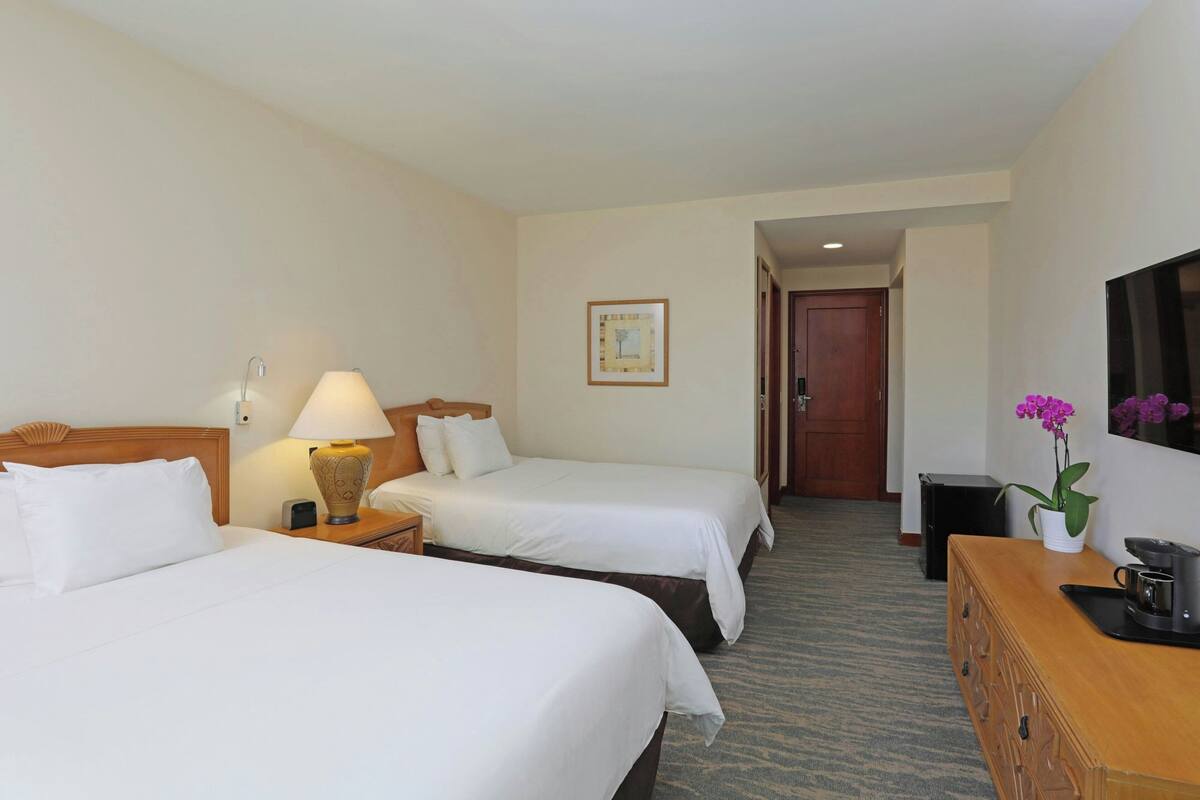 A hotel room with two beds, a desk, a lamp, a coffee maker, and a plant.