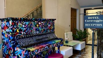 Un'area reception con un pianoforte ricoperto di graffiti, uno sgabello rosa e un cartello con su scritto 'HOTEL Caravaggio RECEPTION'.