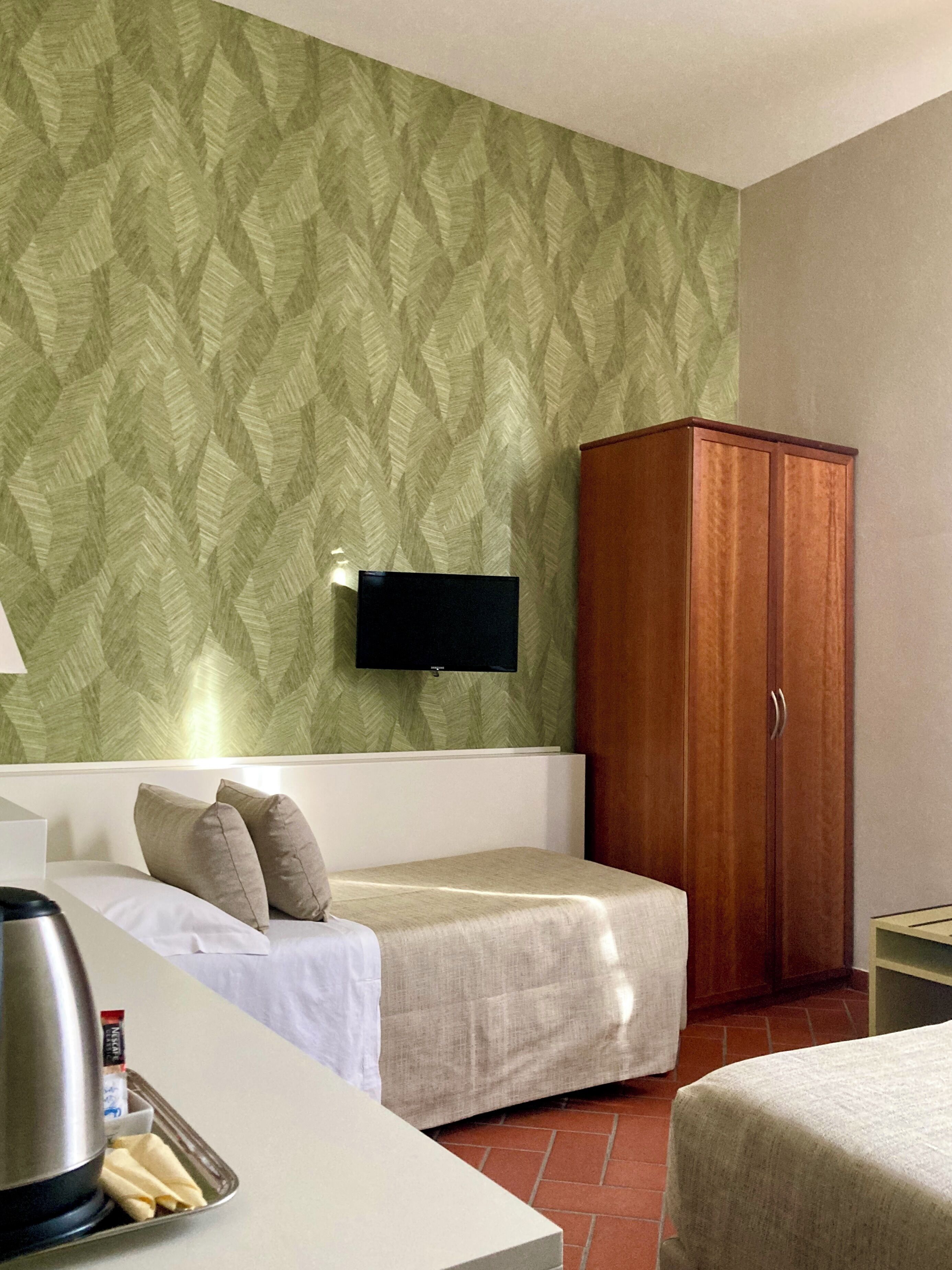 A hotel room with a bed, a wooden wardrobe, a television mounted on the wall, and a small table with a kettle and cups.