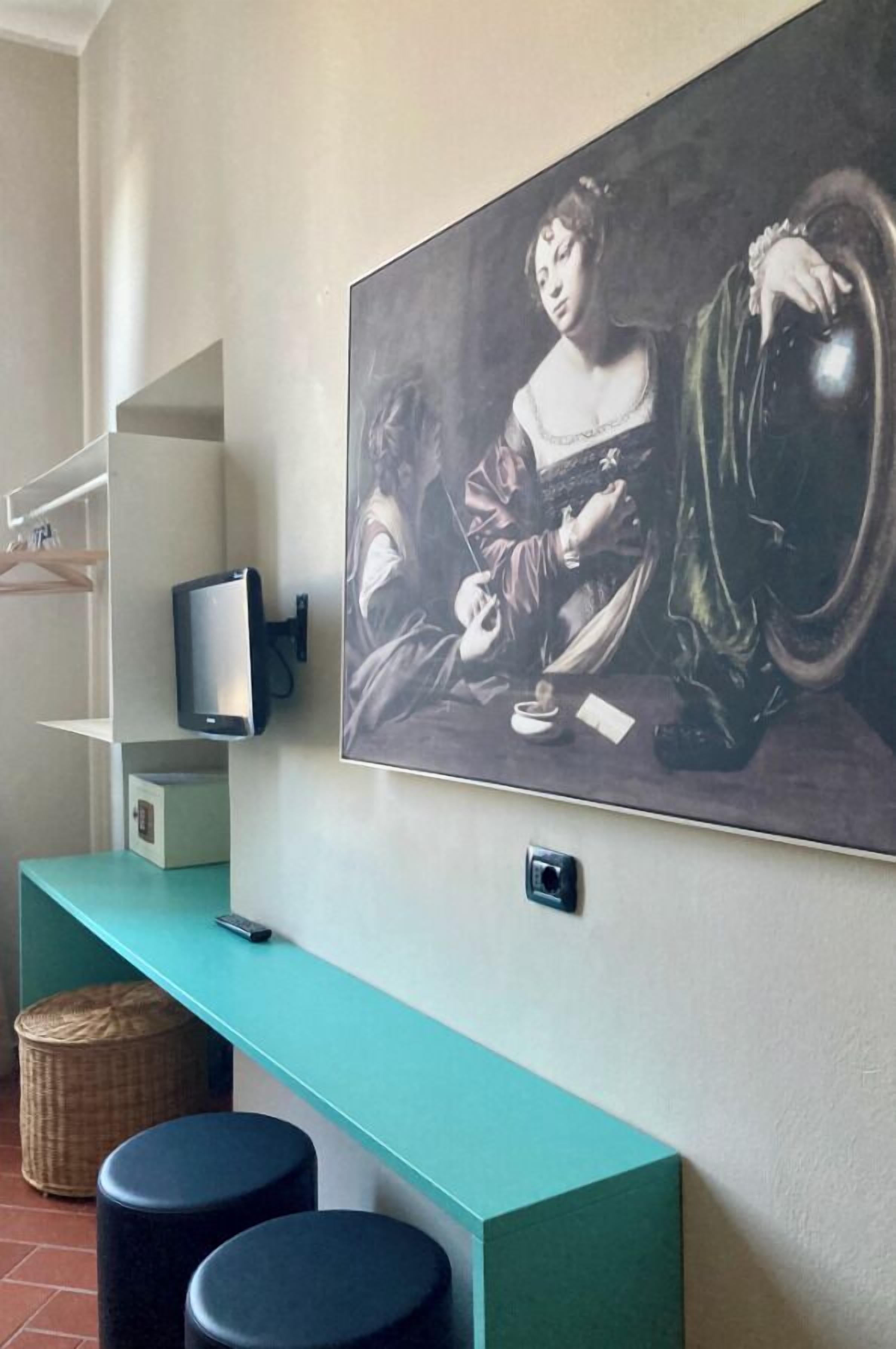 A room with a teal bench, a large painting on the wall, a television mounted on the wall, and a small basket on the floor.