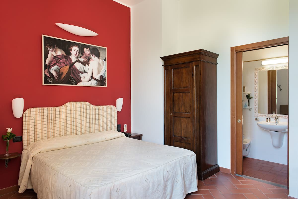 Un dormitorio con cama, un armario de madera, un cuadro en la pared y un baño visible a través de una puerta abierta.