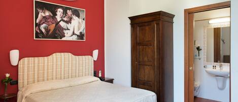 A bedroom with a bed, a wooden wardrobe, a painting on the wall, and a bathroom visible through an open door.