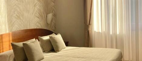 A bedroom with a bed, a headboard, a wall-mounted light, and a window with sheer curtains.