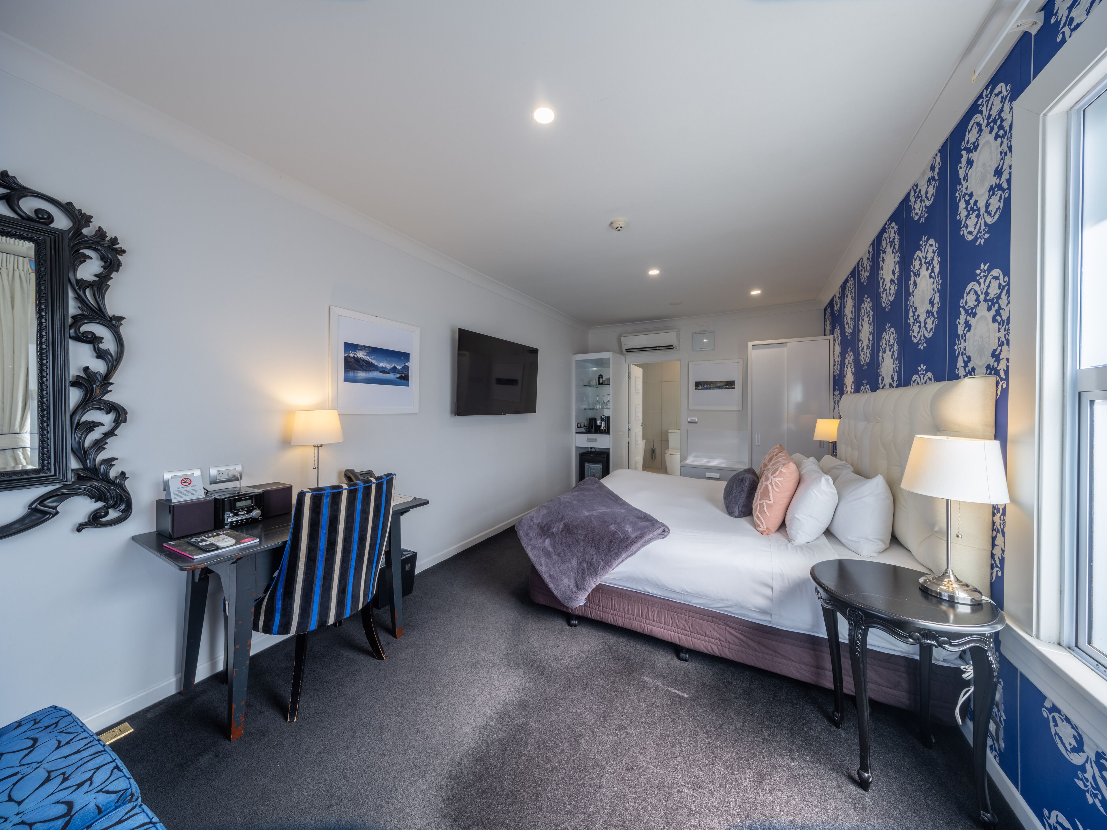 A hotel room with a bed, bedside table, desk, chair, and a large window.