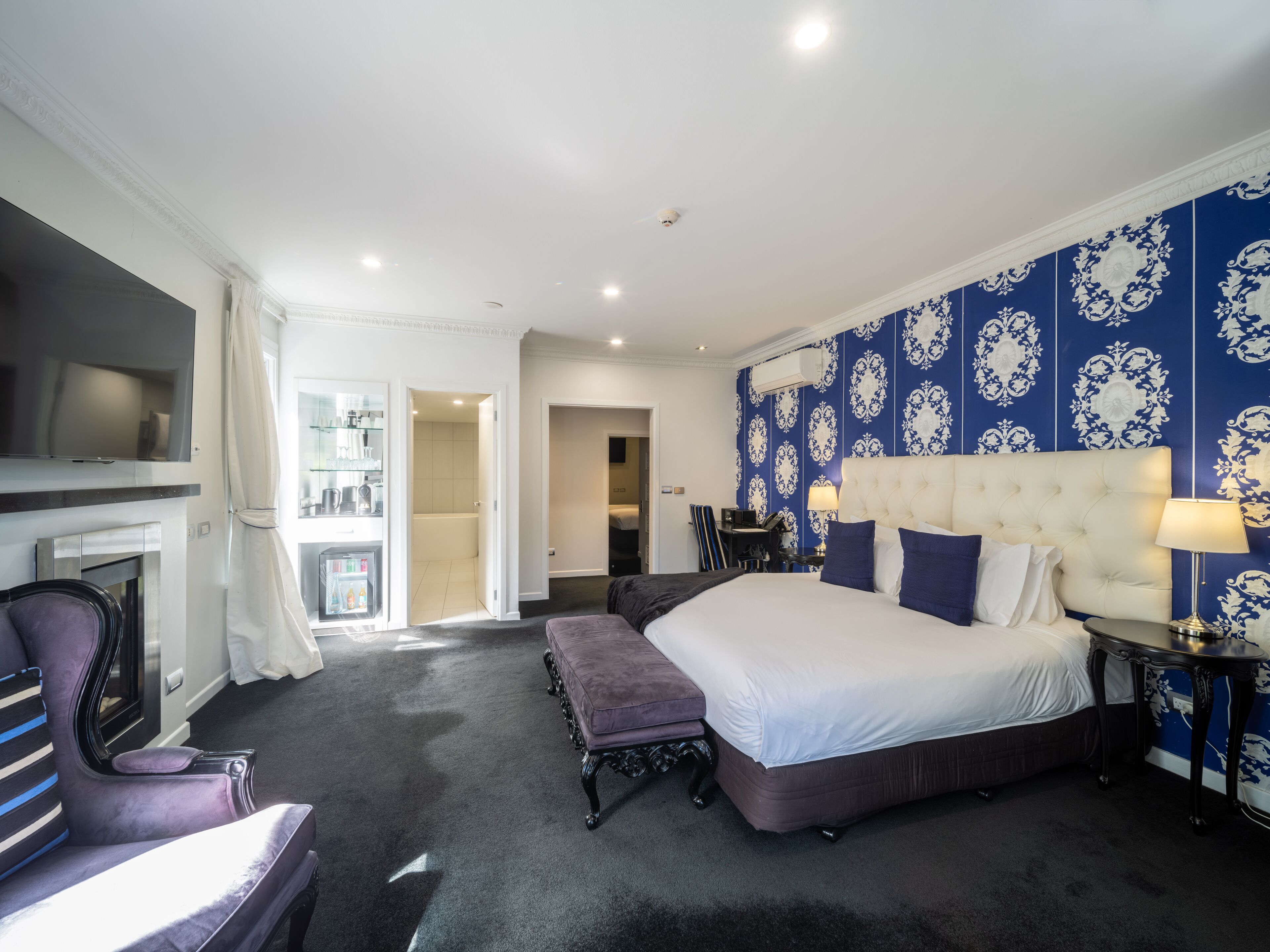 A modern hotel room with a large bed, a TV, a seating area, and a bathroom visible through an open door.