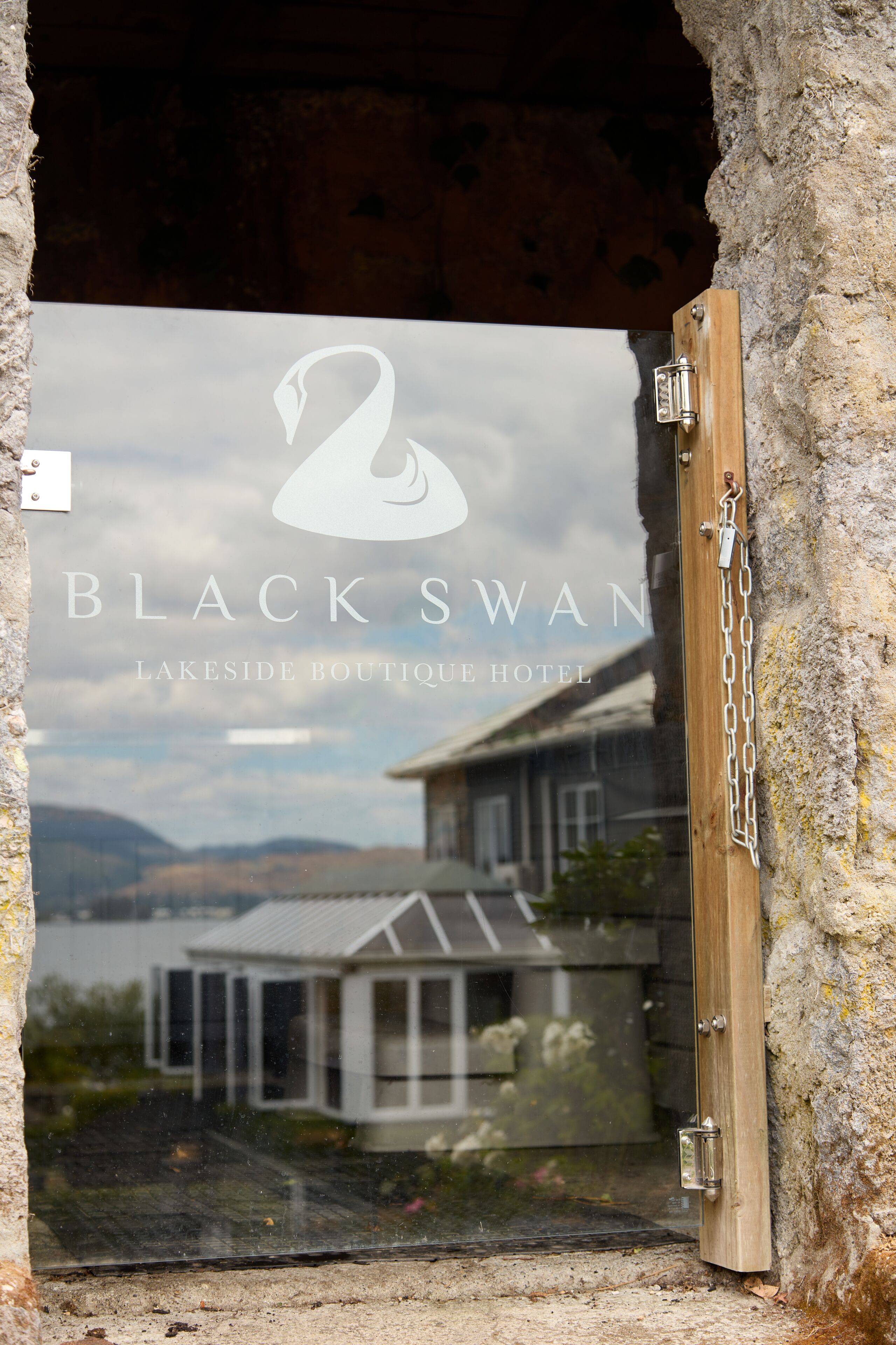 Une porte vitrée ornée du logo du Black Swan Lakeside Boutique Hotel, avec vue sur une maison et un lac en arrière-plan.