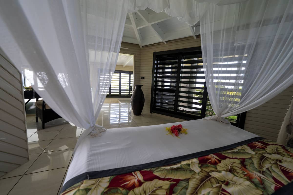 A bedroom with a bed, white curtains, a vase, and a window with blinds.