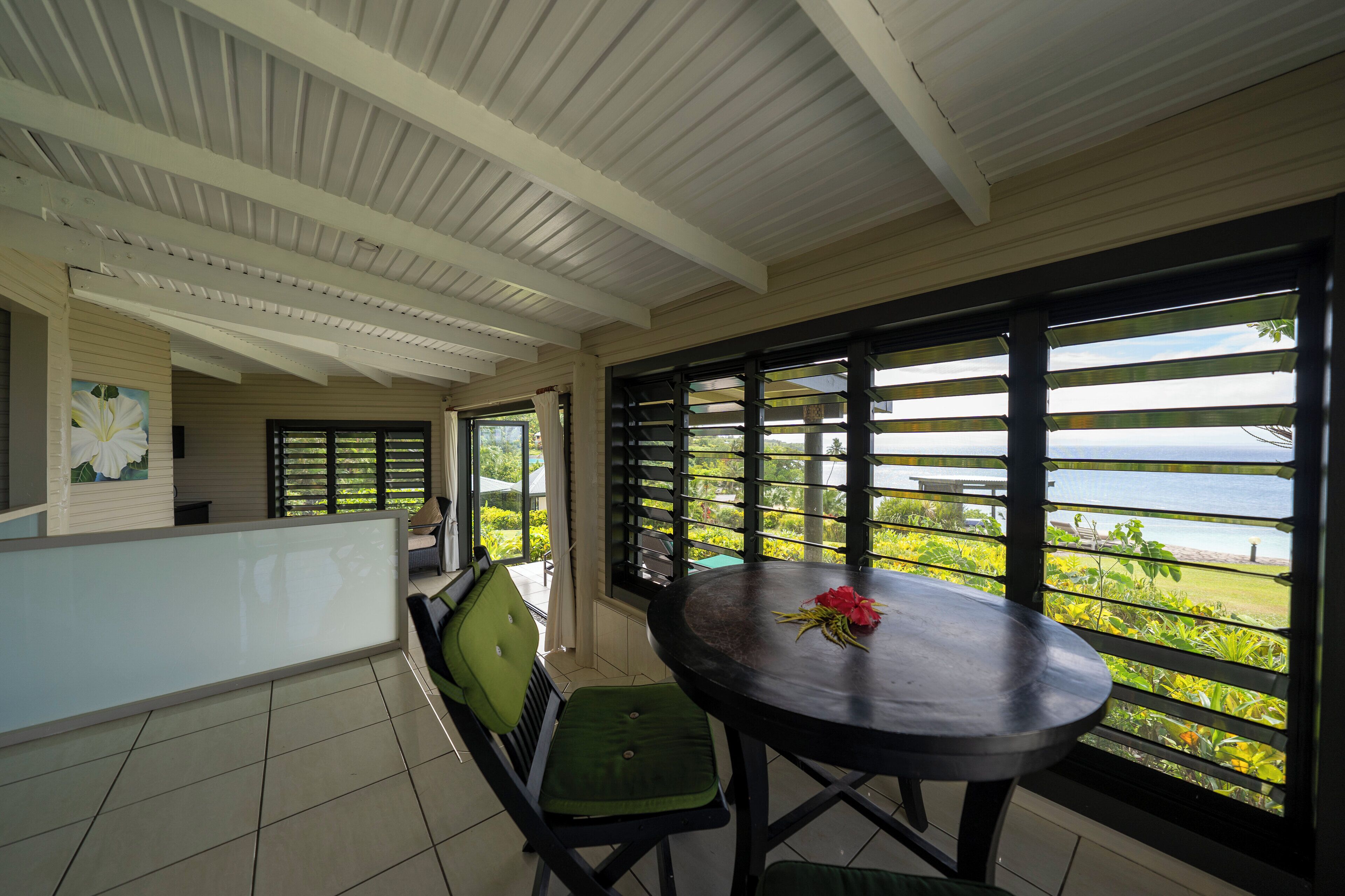 A covered outdoor seating area with a table and chairs, a view of the sea, and a painting on the wall.