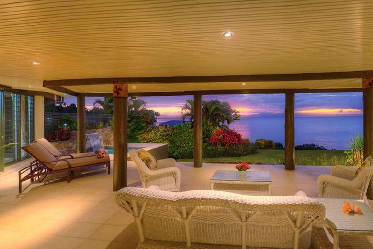 A covered outdoor seating area with wicker furniture, a small table, and a view of the ocean at sunset.