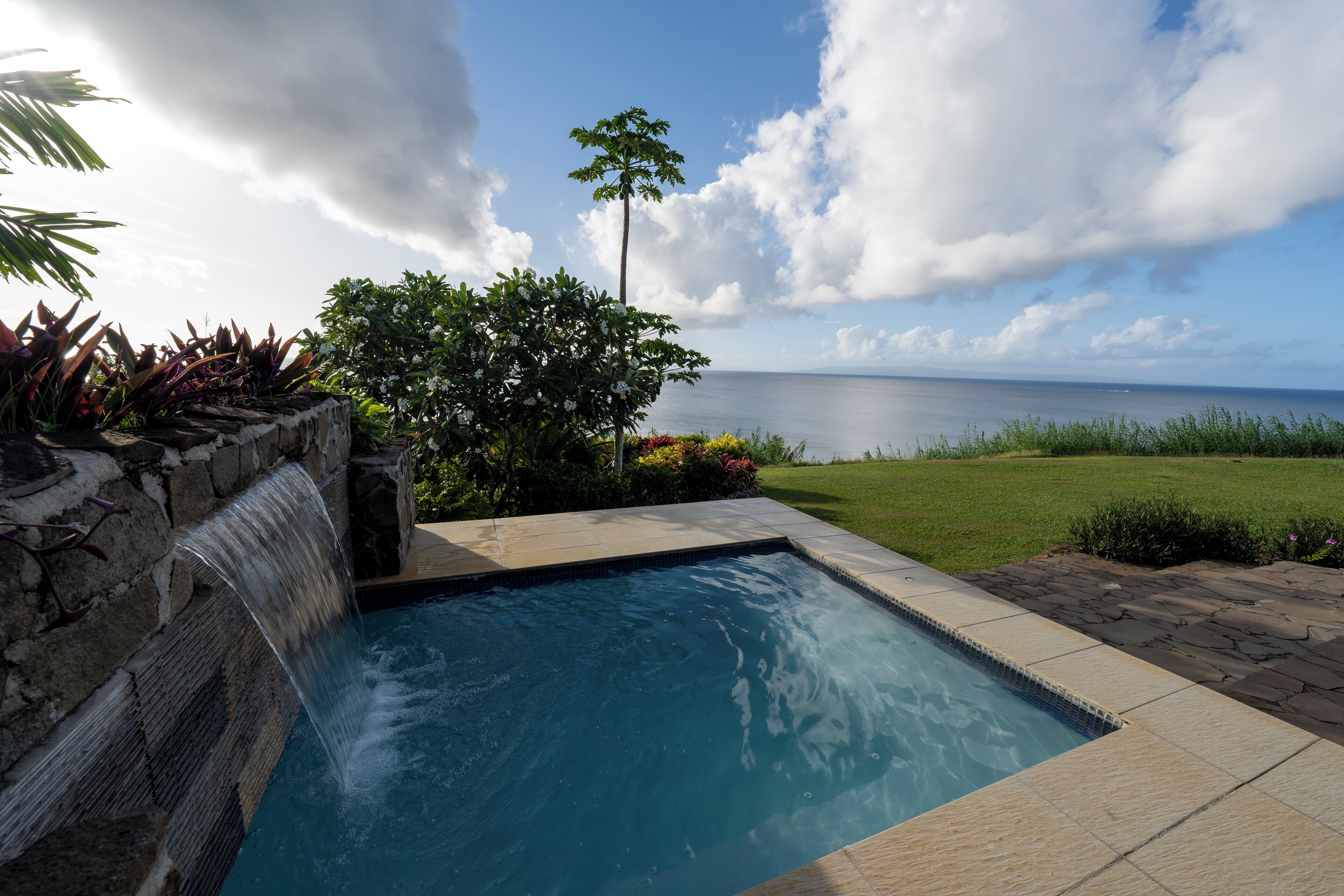 A swimming pool with a waterfall feature, surrounded by a stone wall and lush greenery, overlooking a grassy area and the sea.