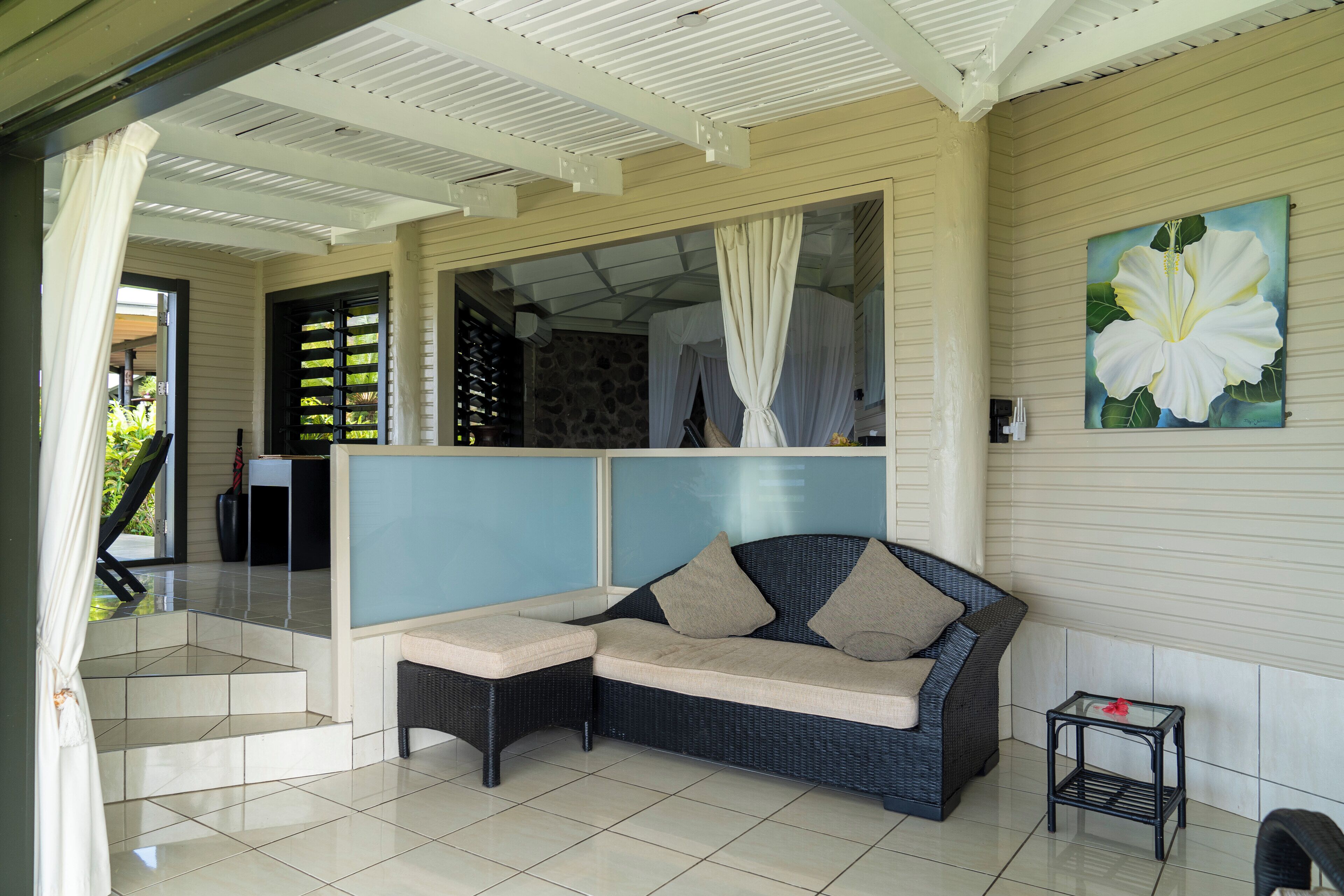 A covered outdoor seating area with a sofa and a small table, a staircase leading to an upper level, and a large flower painting on the wall.