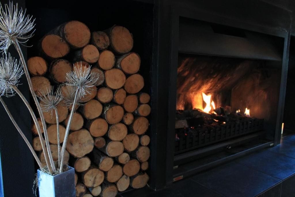 A fireplace with a fire burning, surrounded by neatly stacked logs and decorative dried plants.