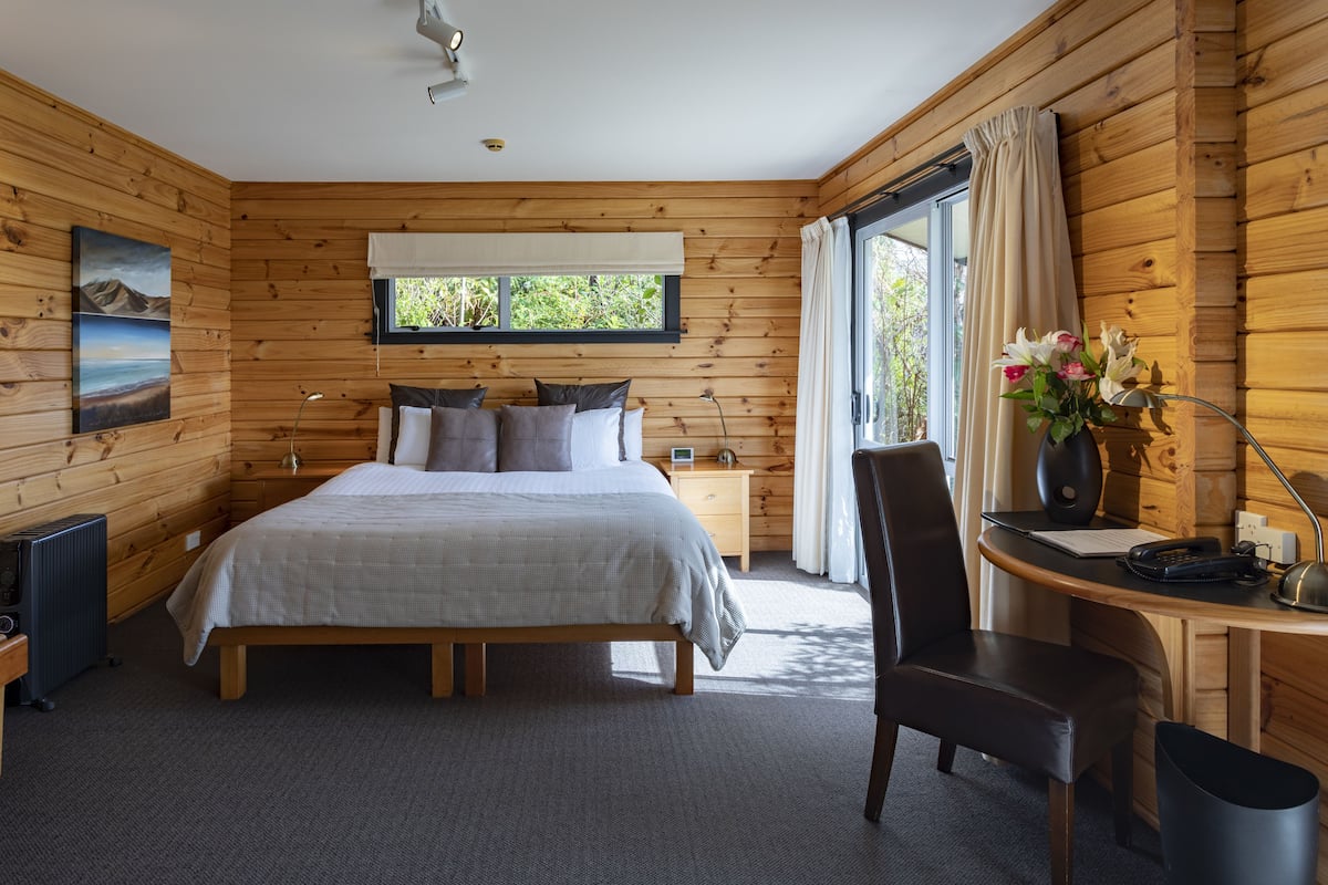 Une chambre avec un grand lit, des murs en bois, un bureau avec une chaise, un radiateur et une fenêtre avec des rideaux.