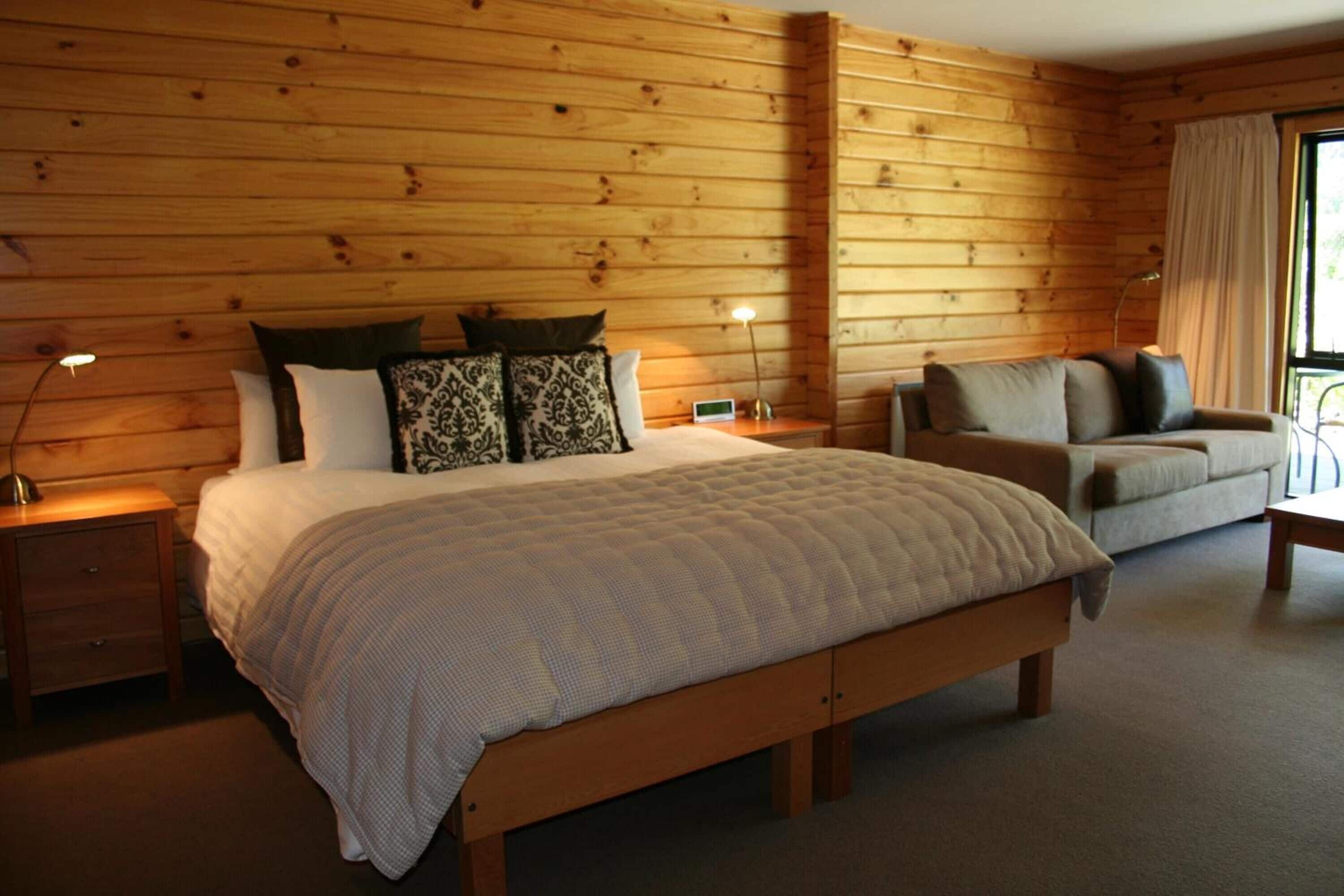 A wooden-paneled room with a bed, a sofa, a nightstand, and a lamp.