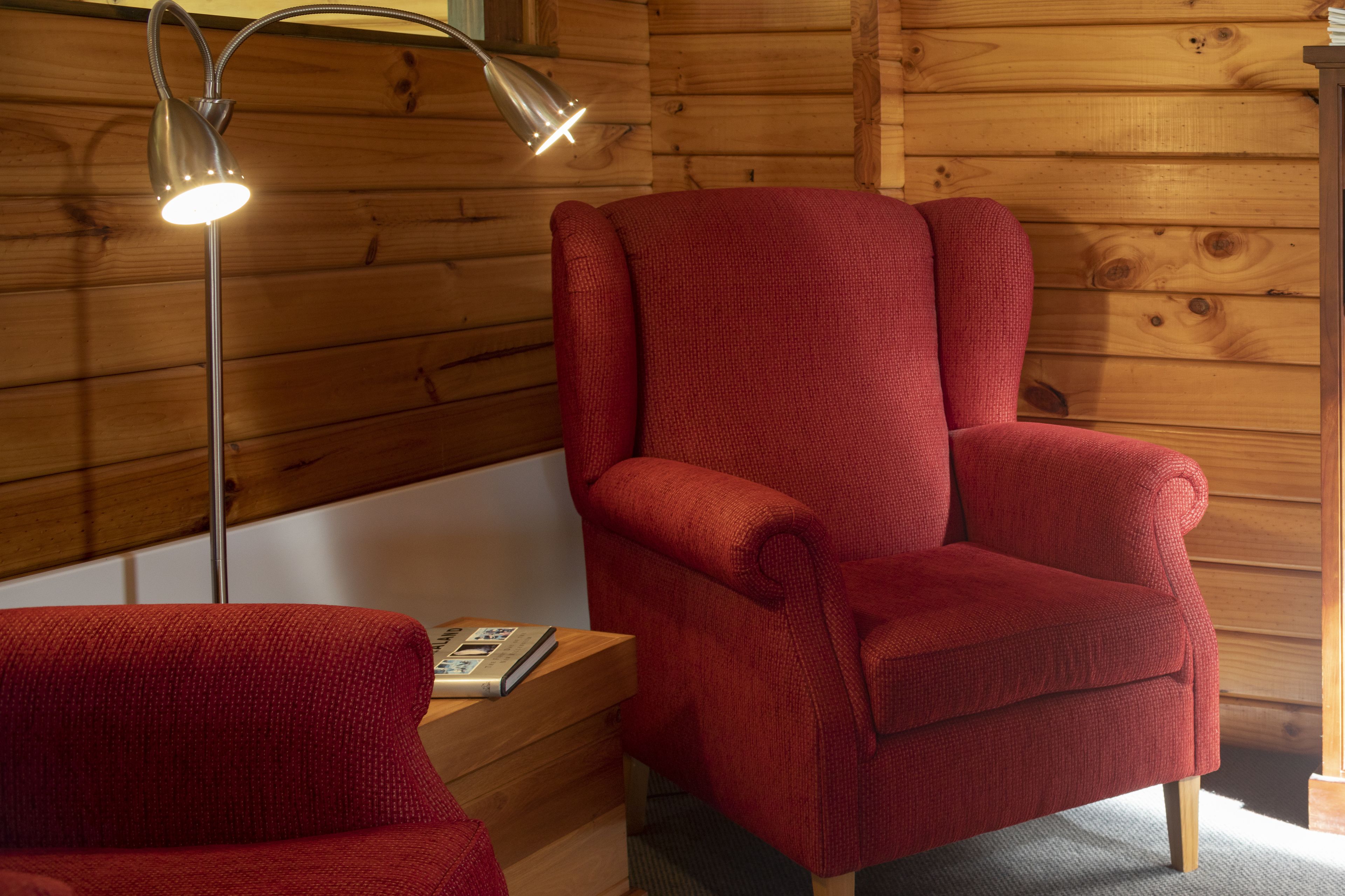 A wooden cabin interior with a red armchair, a floor lamp, and a book on a wooden table.