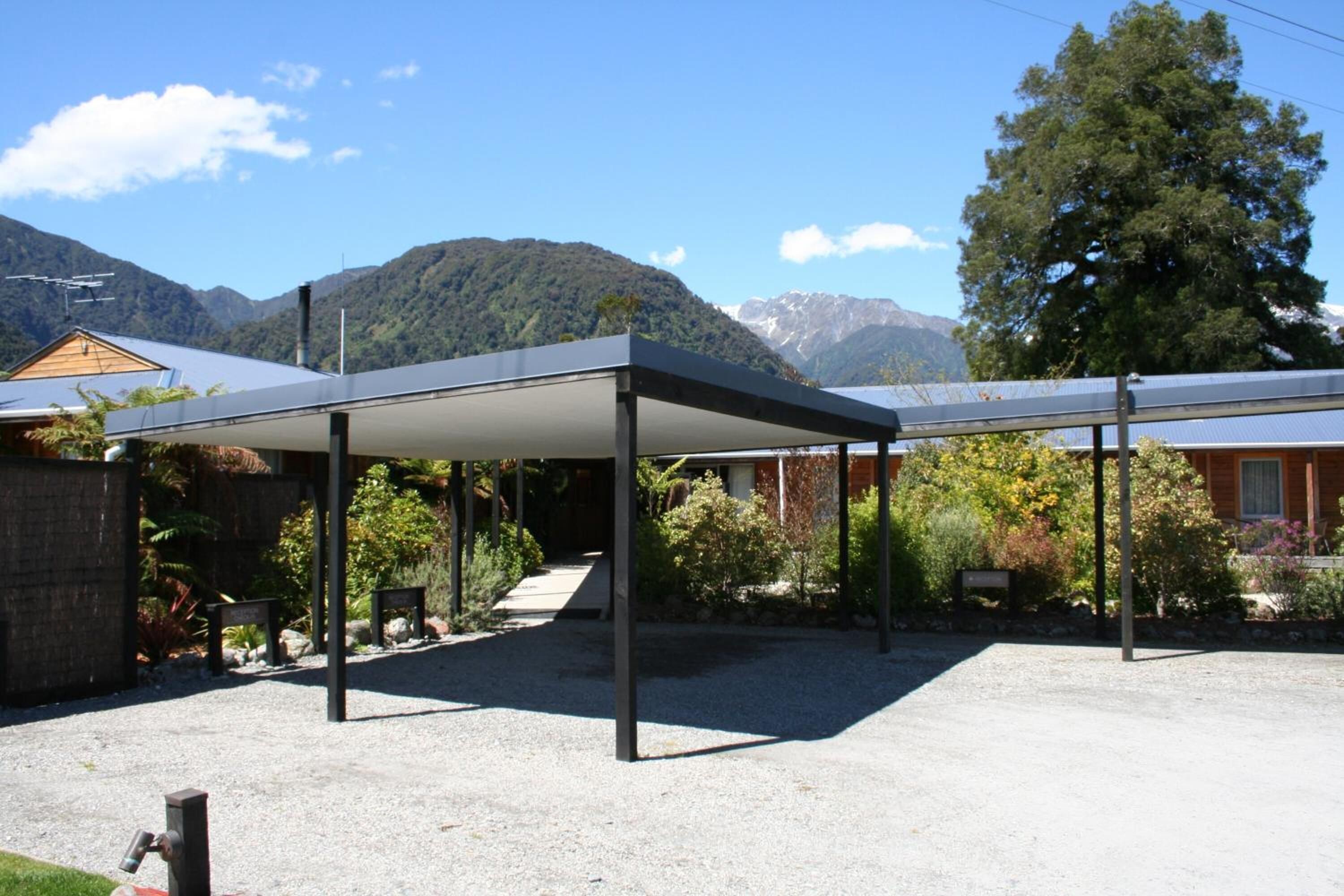 A covered outdoor area with a gravel ground, surrounded by greenery and a mountain backdrop.