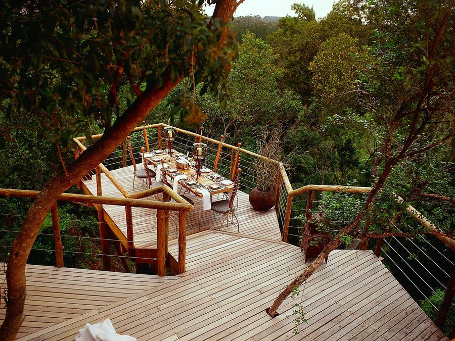 A wooden deck with a dining table set for four, surrounded by chairs, overlooking a forested area.
