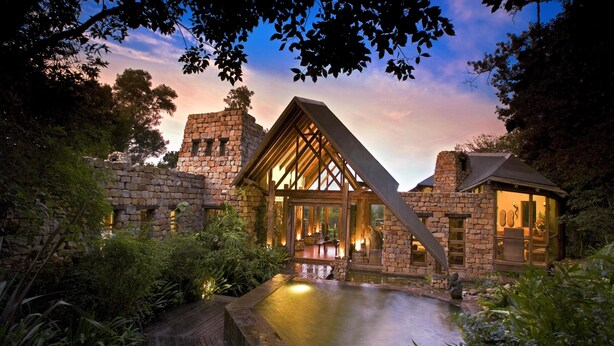 A stone building with a glass facade and a wooden deck leading to a pool.