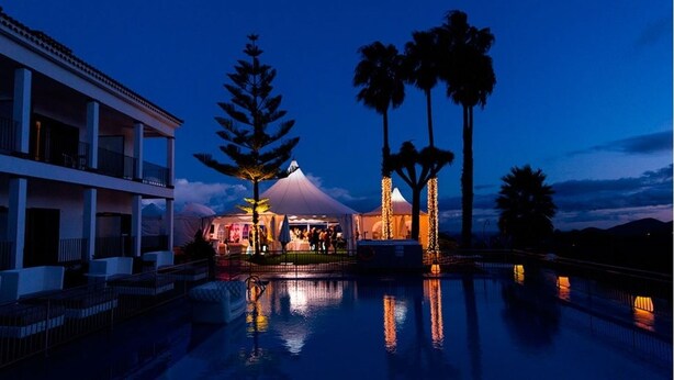 Un resort con piscina, una zona techada con palmeras y un edificio con balcones.