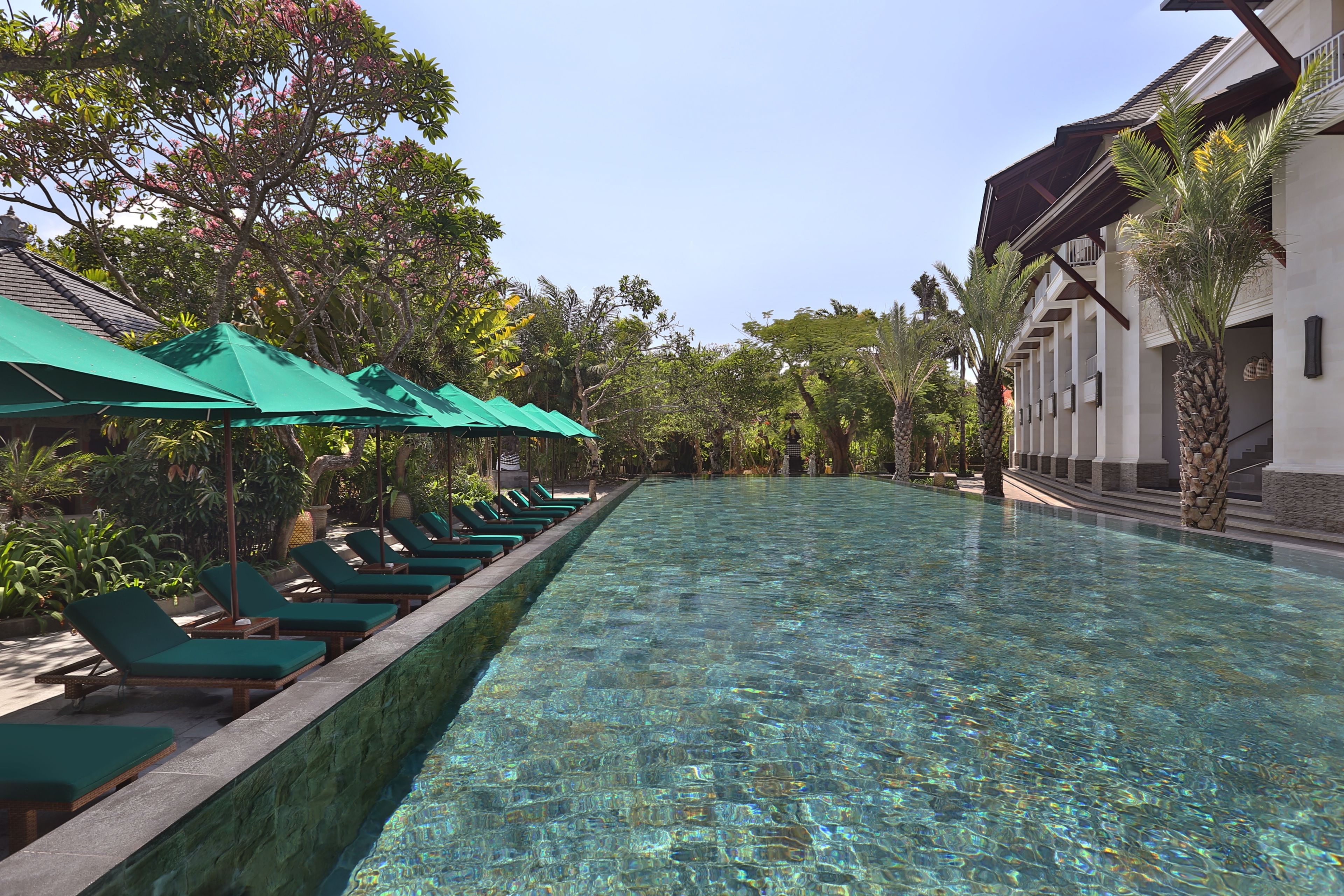 A poolside area with lounge chairs and umbrellas, surrounded by lush greenery and a building with a tiled roof.