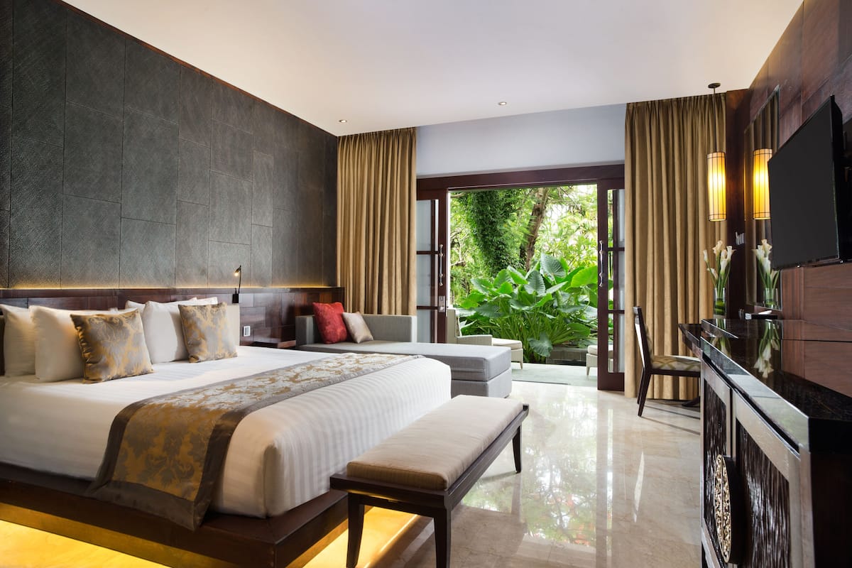 A modern hotel room with a large bed, a desk, a TV, and a view of greenery.