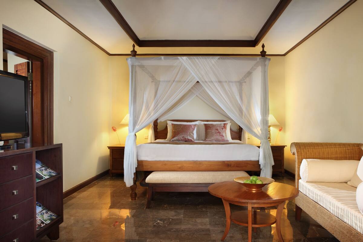 A bedroom with a four-poster bed, a television, a wicker sofa, and a small round table with a bowl.