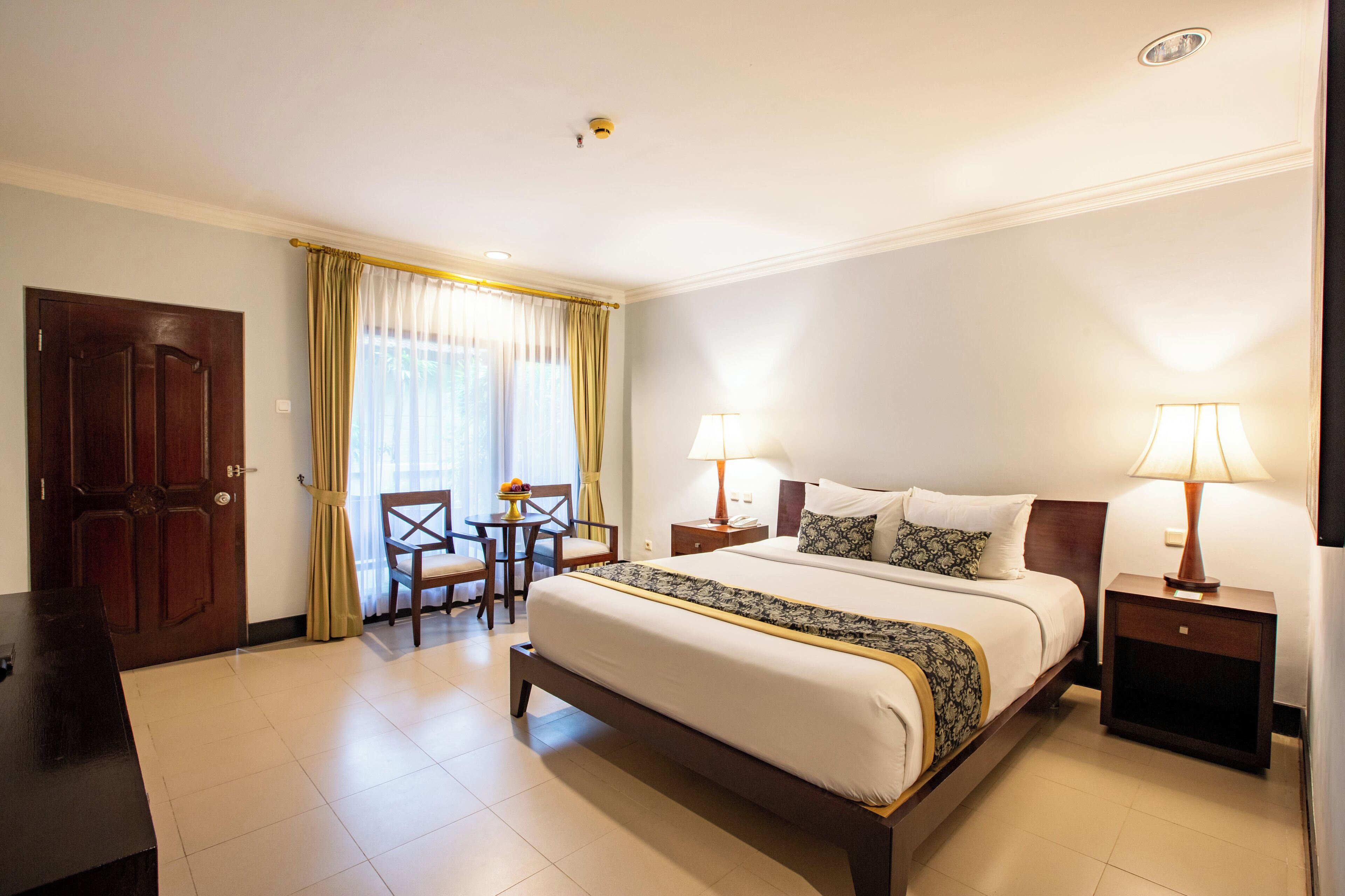 A hotel room with a large bed, two bedside tables with lamps, a wooden door, a small table with chairs, and a window with curtains.