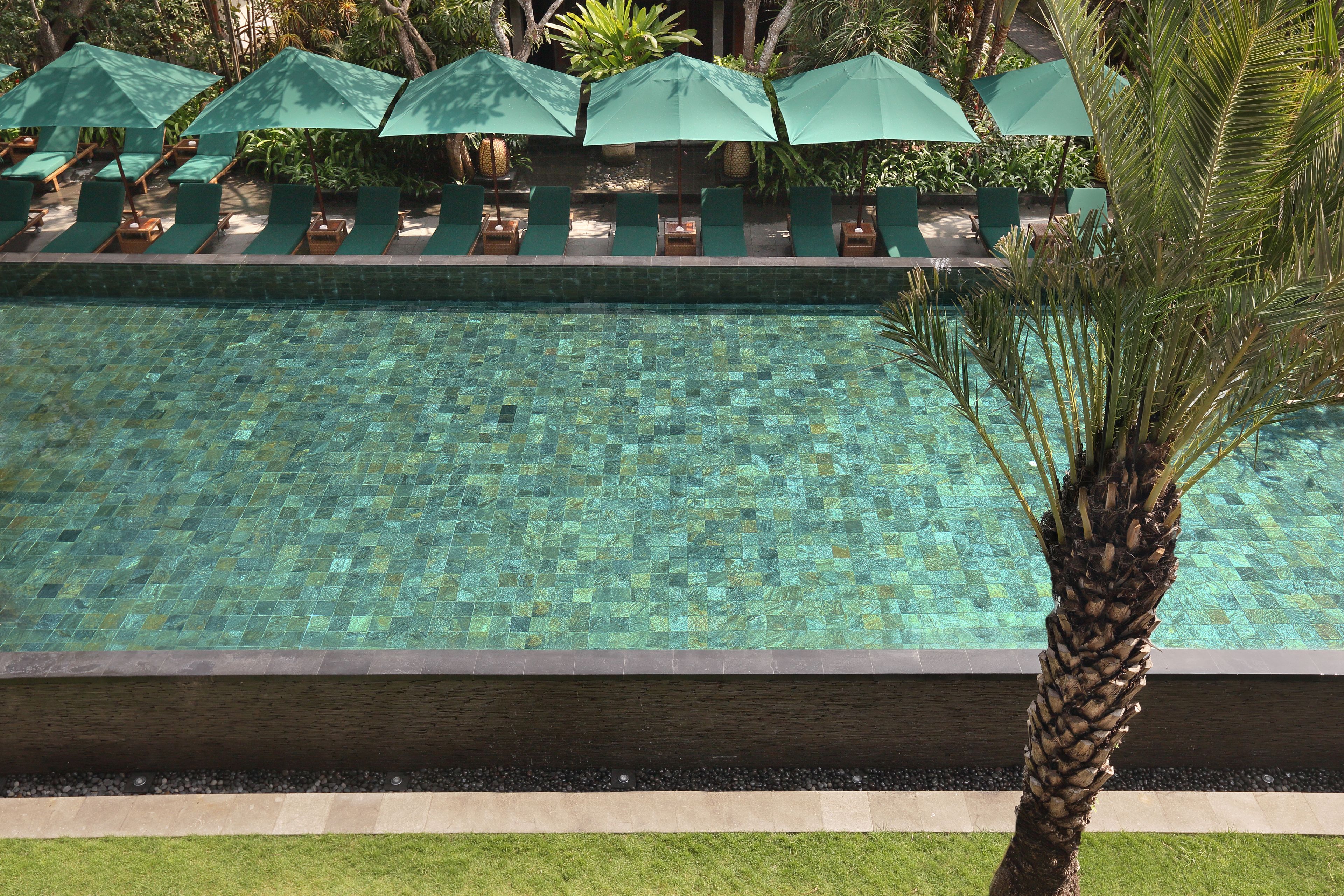 Una piscina con superficie de baldosas, rodeada de sillas de descanso y sombrillas verdes.