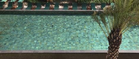 Una piscina con superficie de baldosas, rodeada de sillas de descanso y sombrillas verdes.