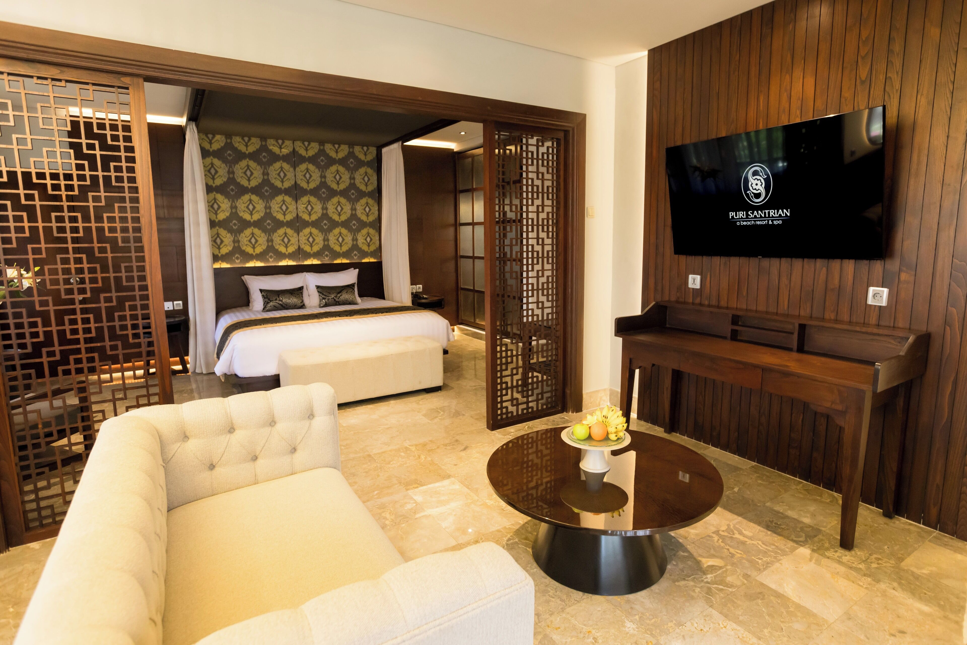 A hotel room with a bed, a TV, a sofa, a coffee table with fruit, and a wooden partition.
