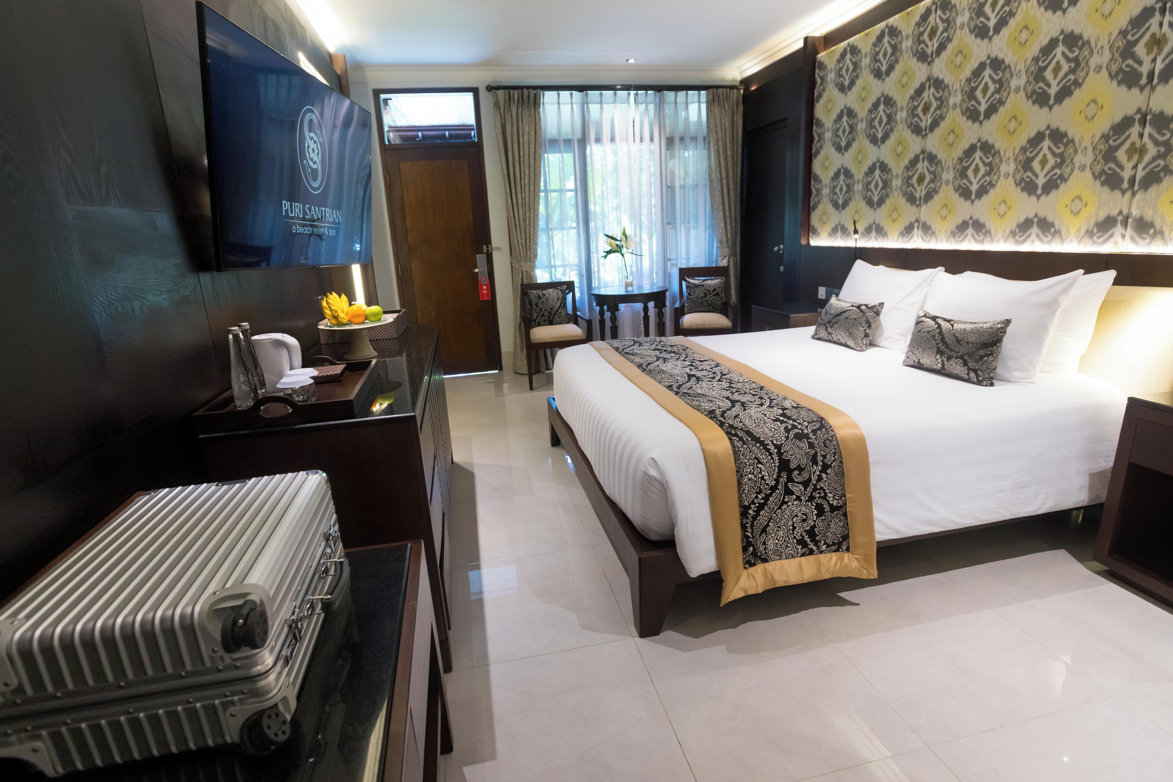 A hotel room with a bed, a desk with a computer, a chair, a TV, and a suitcase.