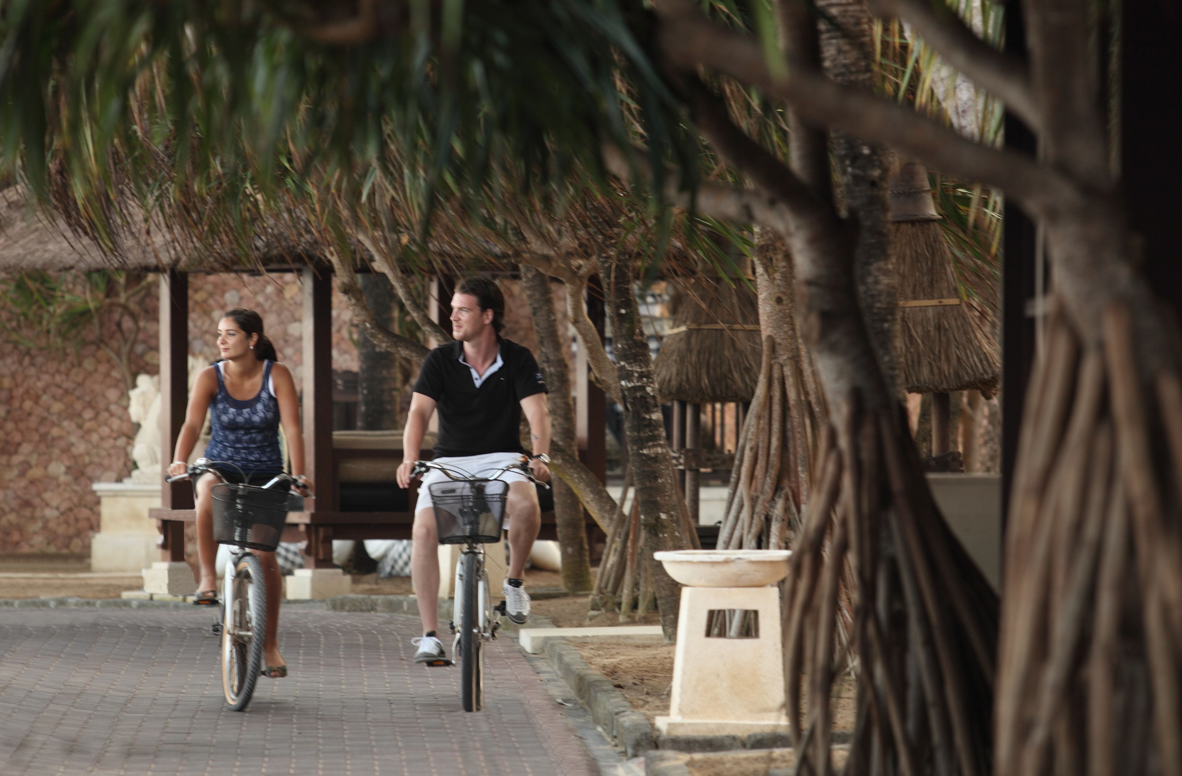 Dos personas montando en bicicleta por un camino pavimentado, con palmeras y una estructura de piedra al fondo.