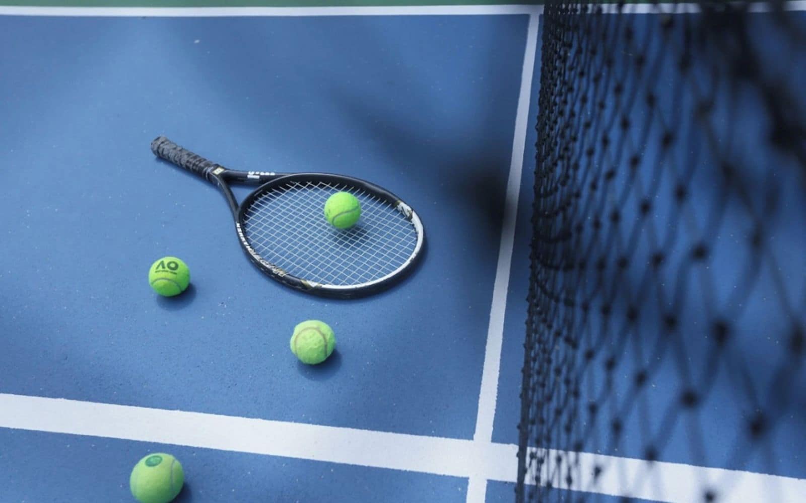 A tennis court with a net, a tennis racket, and three tennis balls.