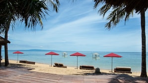 Ein Strand mit Holzterrasse, roten Sonnenschirmen und Liegestühlen.