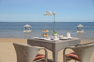 A beachside dining table with wicker chairs, a flower arrangement, and a three-tiered fruit and pastry display.