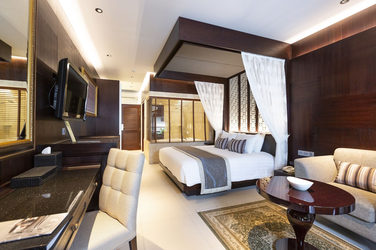 A modern hotel room with a large bed, a sitting area with a sofa, a dining table, and a television mounted on the wall.