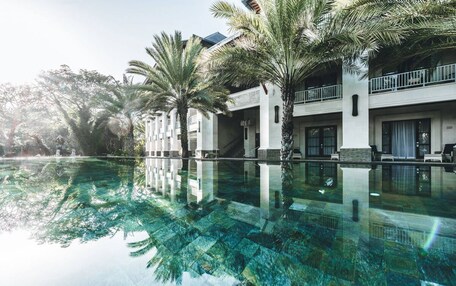A hotel with a large swimming pool surrounded by palm trees, reflecting the architecture and greenery.