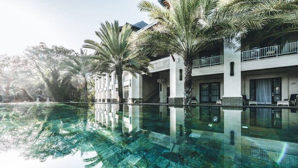 A hotel with a large swimming pool surrounded by palm trees, reflecting the architecture and greenery. - Puri Santrian (Denpasar)