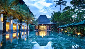A pool area with a reflection of a building and palm trees, surrounded by lush greenery.