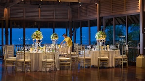 Ein Festsaal mit runden Tischen, gedeckt für eine formelle Veranstaltung, große Fenster mit Blick aufs Meer und eine hohe Decke mit Holzbalken.