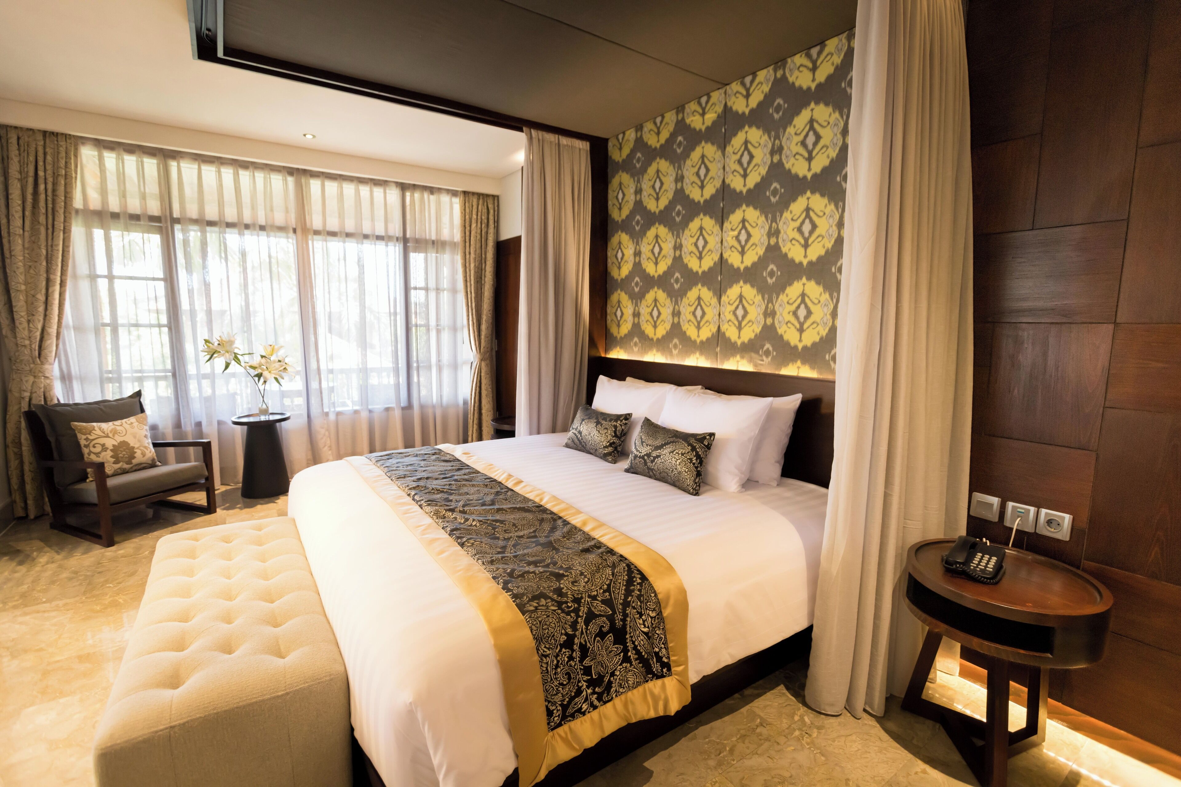 Quarto de hotel com uma cama grande, mesa de cabeceira com telefone, uma cadeira e uma pequena mesa com um vaso de flores.