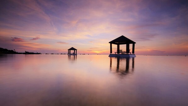 A tranquil sunset over a calm sea with two gazebos on stilts, one with a table and chairs, and the other empty. - Puri Santrian (Denpasar)