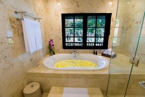 A bathroom with a large bathtub filled with yellow flowers, a glass shower enclosure, and a window with a view of greenery.