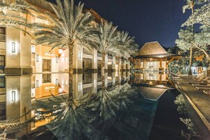 A nighttime view of a hotel with a pool reflecting the building and palm trees.