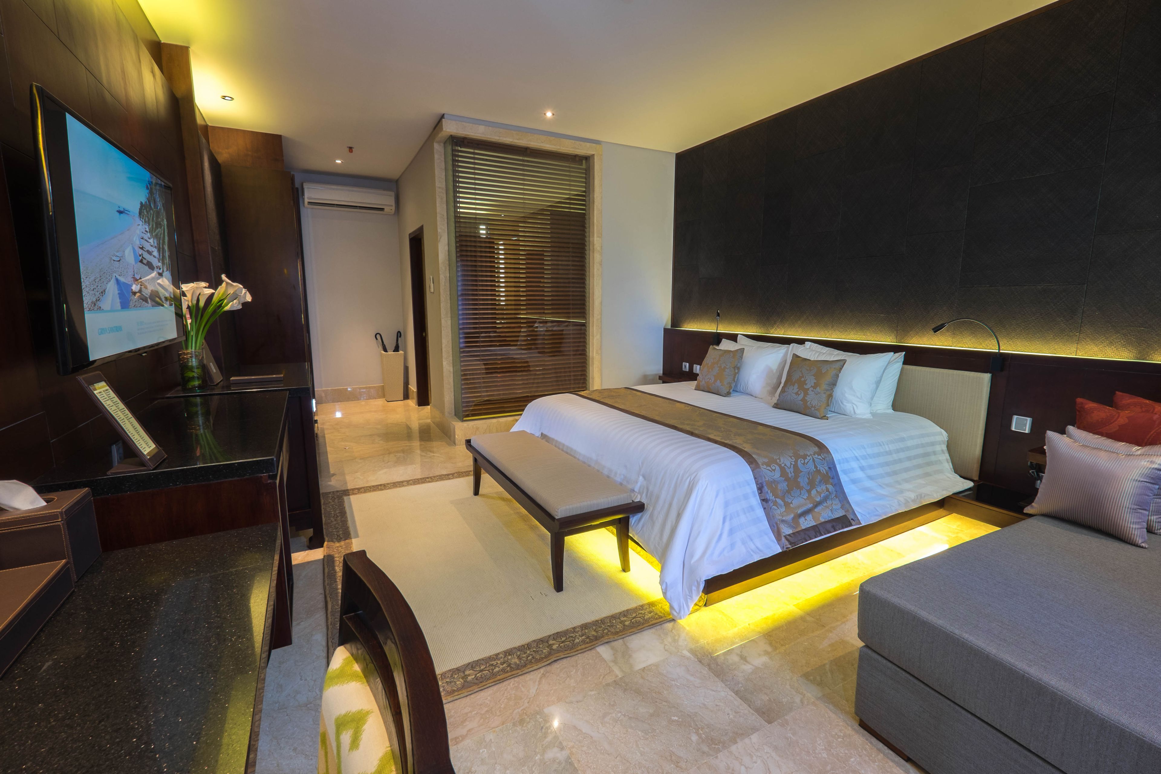 A modern hotel room with a large bed, a desk with a computer, a bench, and a TV on the wall.