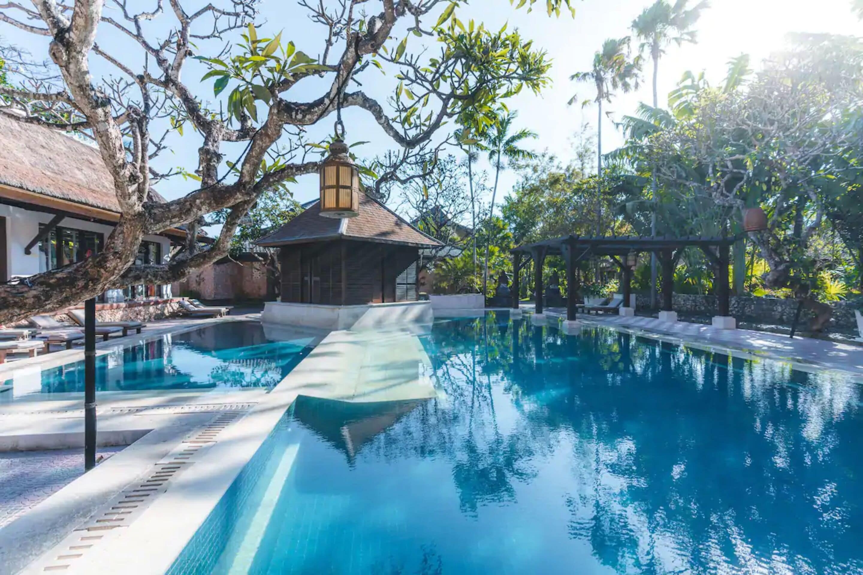 A clear swimming pool with lounge chairs and a gazebo, surrounded by trees and a building with a thatched roof.