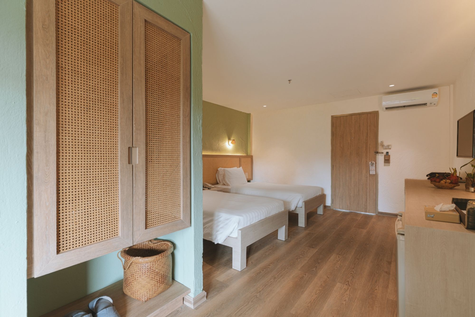 Habitación de hotel con dos camas, un armario de madera, una cesta de mimbre y un pequeño escritorio con un frutero.