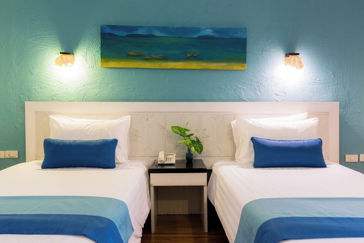 Een hotelkamer met twee bedden, een nachtkastje met een telefoon, een schilderij van boten op het water en wandlampen.